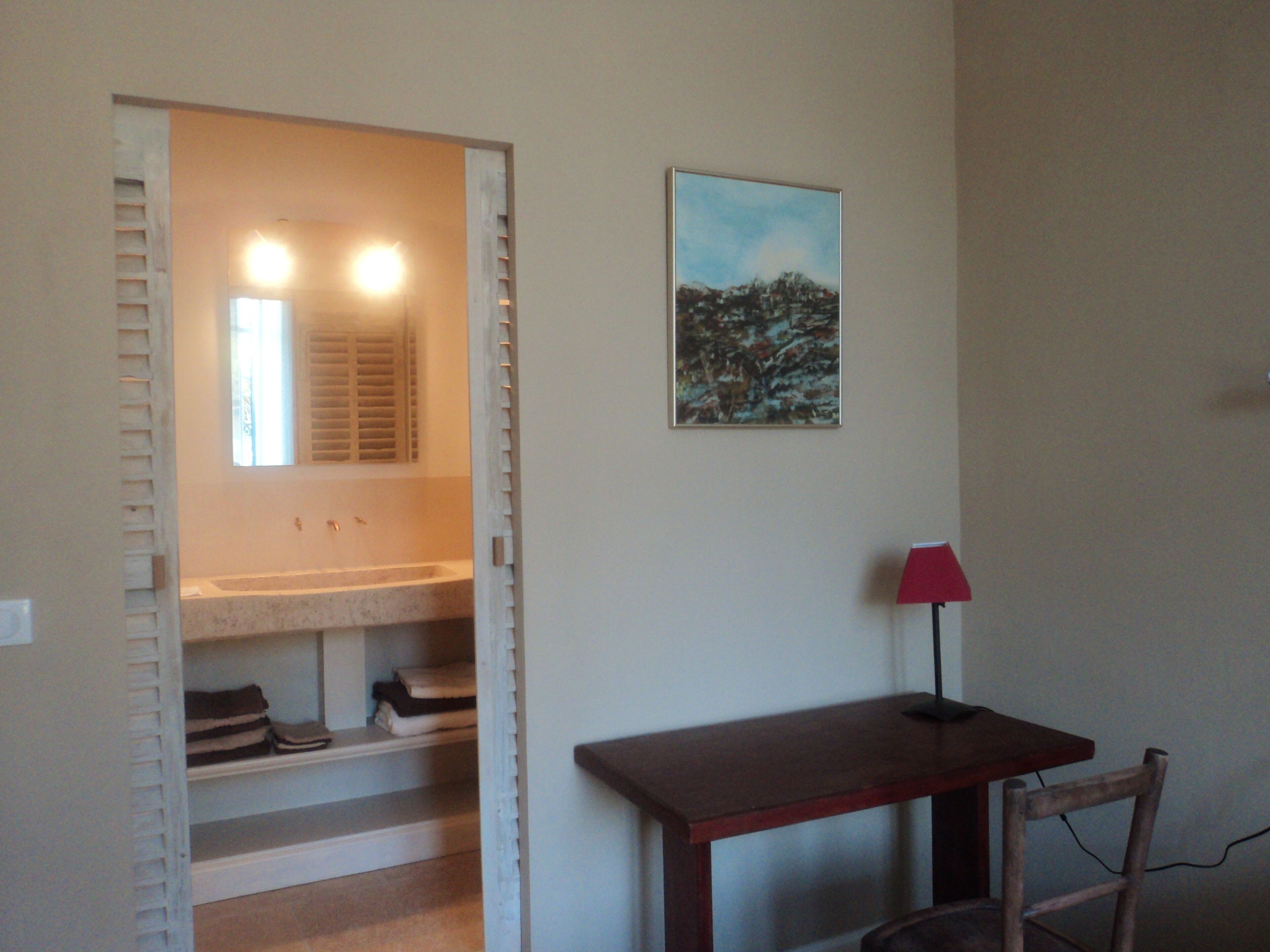 A bathroom with a shower, a sink, and a mirror. There is a painting on the wall and a red lamp on a table.