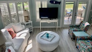 A living room with a white sofa, a round coffee table, a colorful patterned chair, a television on a white stand, and a ceiling fan.