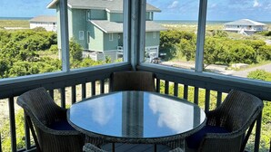A covered outdoor area with a round table and wicker chairs, overlooking a beach and buildings. - NOW AVAILABLE LABOR DAY, OCEAN views, 2 KING masters. 2 clubs- separate purchase (Bald Head Island)
