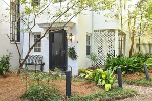 A white building with a black door, a bench, and a garden with plants and a pathway. - The Coach House Circa 1755 (Charleston)