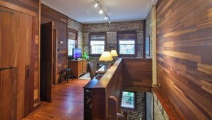 A loft-style room with exposed brick walls, wooden floors, and a long wooden bench. There is a desk with a computer and a lamp, and a window providing natural light. - The Coach House Circa 1755 (Charleston)