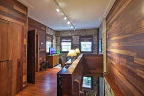 A loft-style room with exposed brick walls, wooden floors, and a long wooden bench. There is a desk with a computer and a lamp, and a window providing natural light.