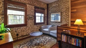 A room with exposed brick walls, a round table, two armchairs, a bookshelf, and a lamp.