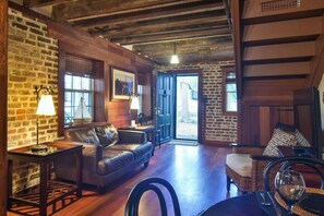 A rustic interior with exposed wooden beams, a leather sofa, wicker furniture, and a dining area with a table and chairs. - The Coach House Circa 1755 (Charleston)