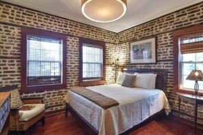 A bedroom with a large bed, two windows with blinds, a wooden headboard, a wicker chair, and a lamp on a side table. - The Coach House Circa 1755 (Charleston)