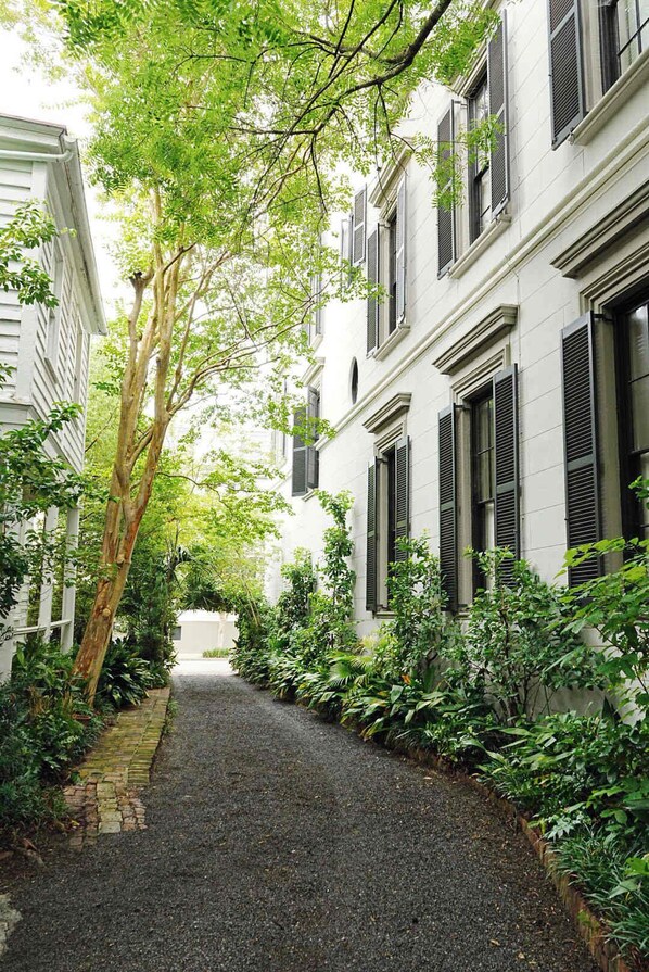 A narrow alleyway flanked by white buildings with black shutters, surrounded by lush greenery and a gravel path. - The Coach House Circa 1755 (Charleston)