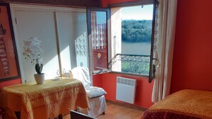 A room with two beds, a table with a chair, a potted plant, and a view of a forest through a window. - Country cottage "Le Romarin" (Lagorce)