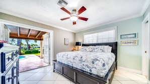 Un dormitorio con cama, ventilador de techo, ventana con persianas y vista a una piscina y vegetación.