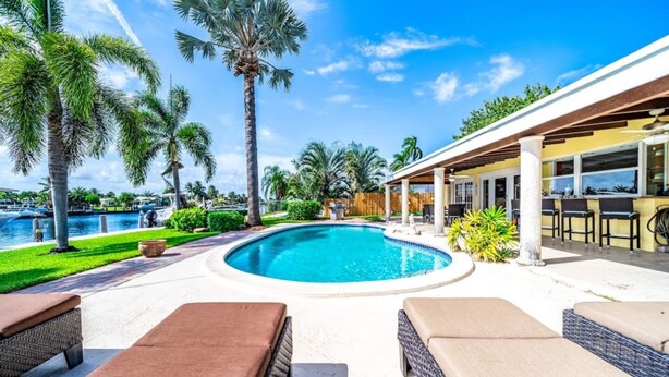Un área de piscina con sillones, un porche cubierto y palmeras.