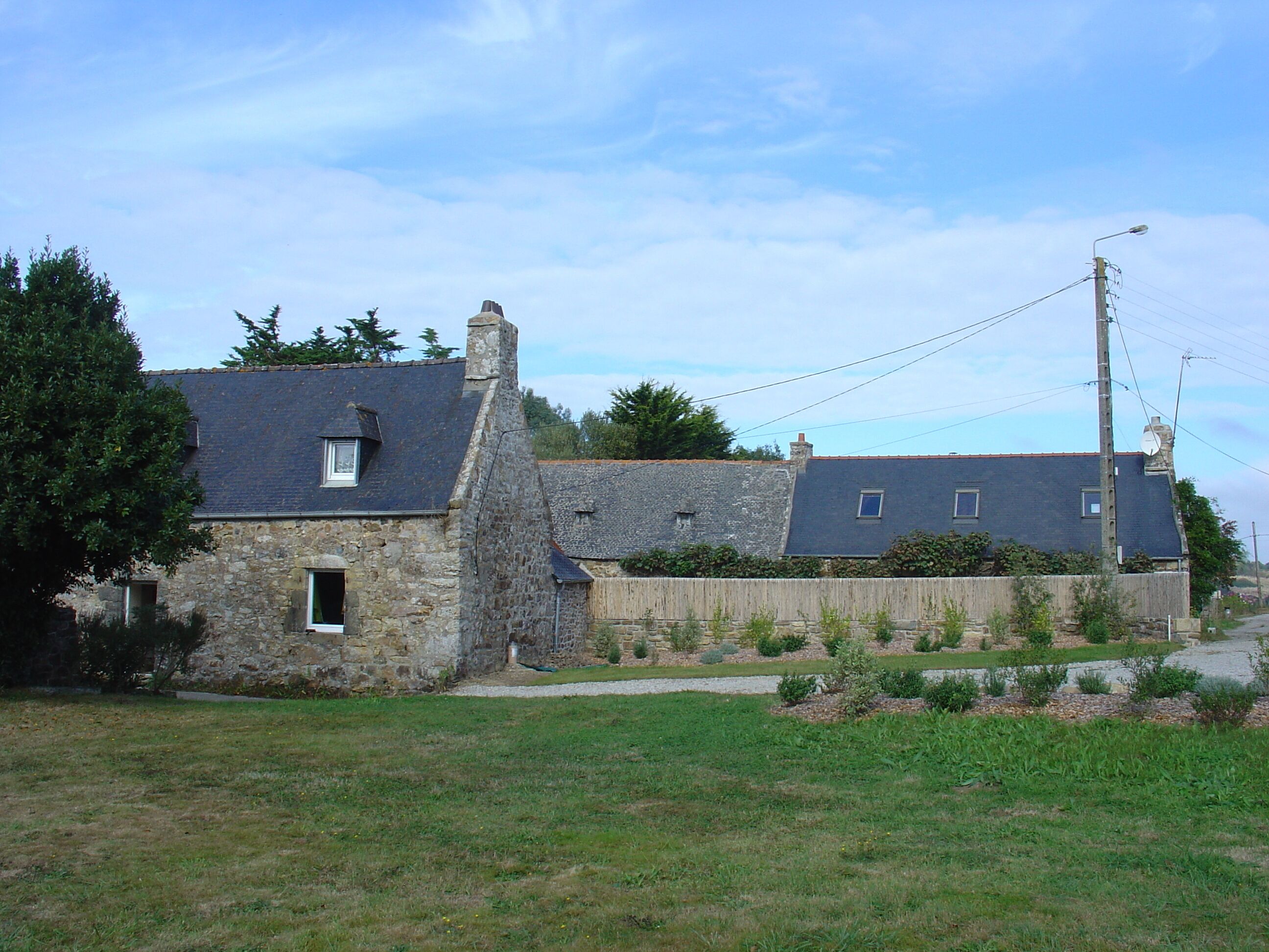 A stone house with a grey roof, a small window, and a garden in front.