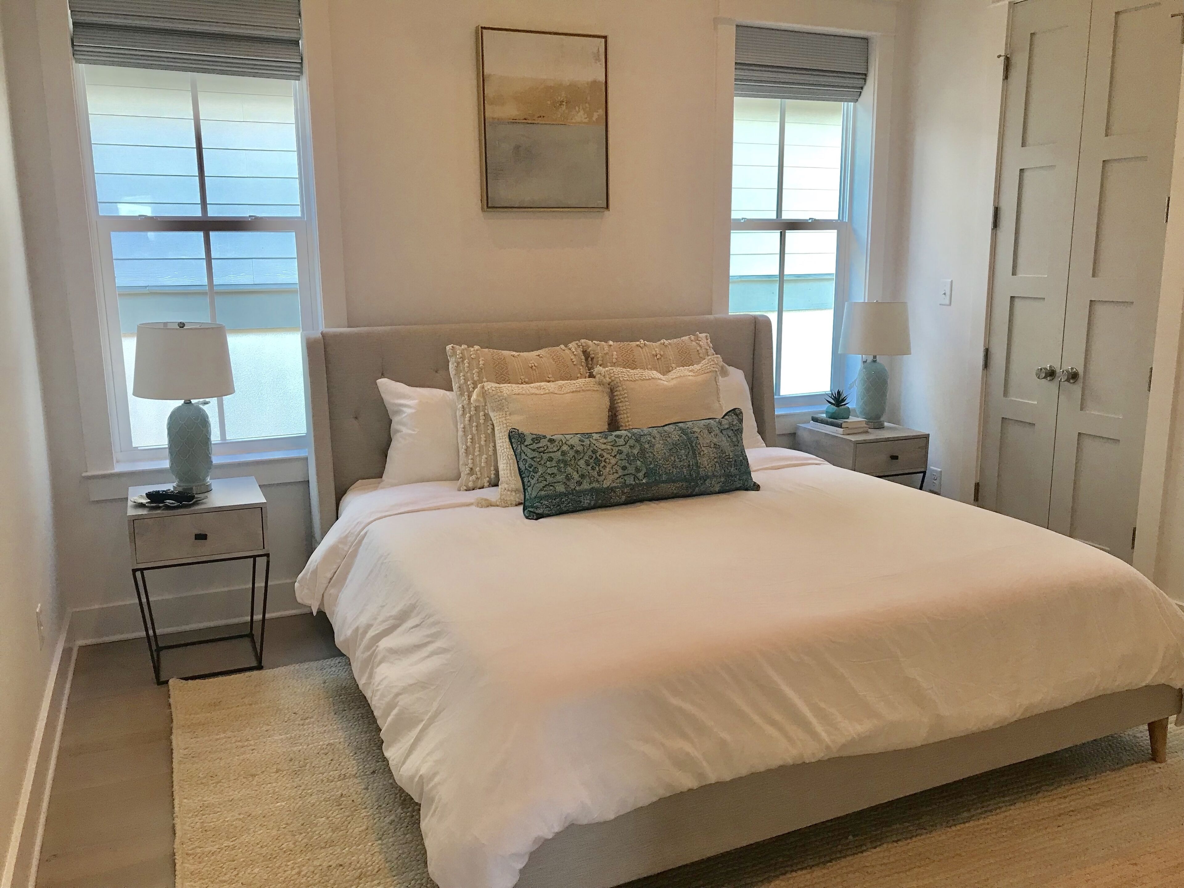 Une chambre à coucher bien rangée, avec un grand lit, deux tables de chevet, une commode, une porte et une fenêtre avec des stores.