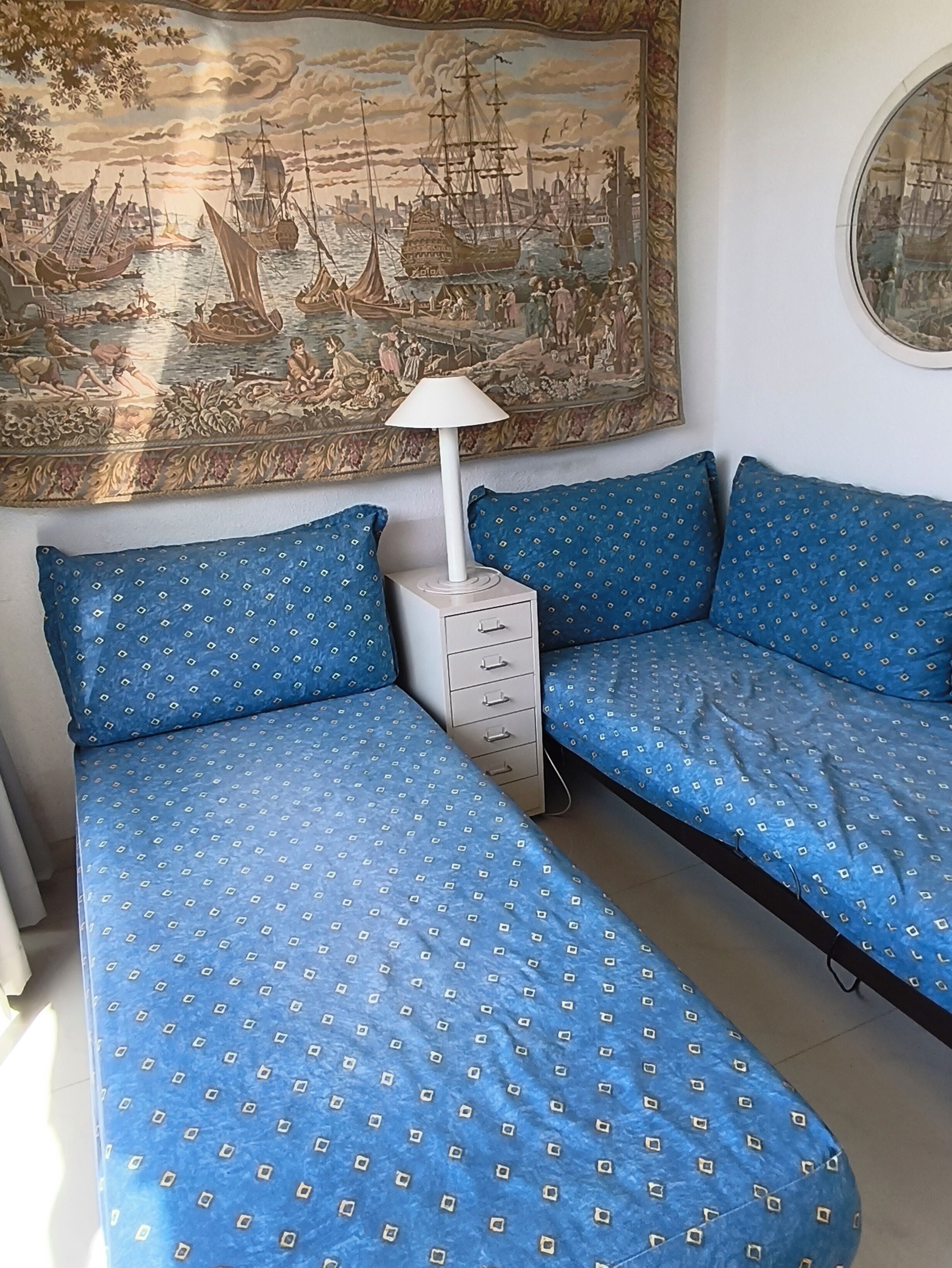 Une chambre comprenant un lit, une table de chevet, une lampe et un grand tableau représentant un paysage portuaire.
