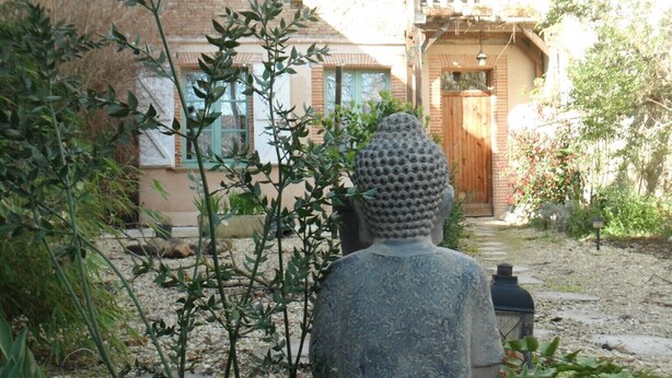 A garden with a stone Buddha statue, surrounded by greenery and a pathway leading to a building with a wooden door and windows. - Quiet, restful gîte in town. Everything is less than 500m away on foot! (Beaumont-De-Lomagne)