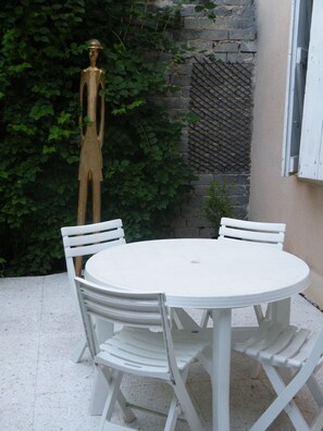 A round white table with four chairs, a white window, and a wall with a metal grid and greenery. - Quiet, restful gîte in town. Everything is less than 500m away on foot! (Beaumont-De-Lomagne)