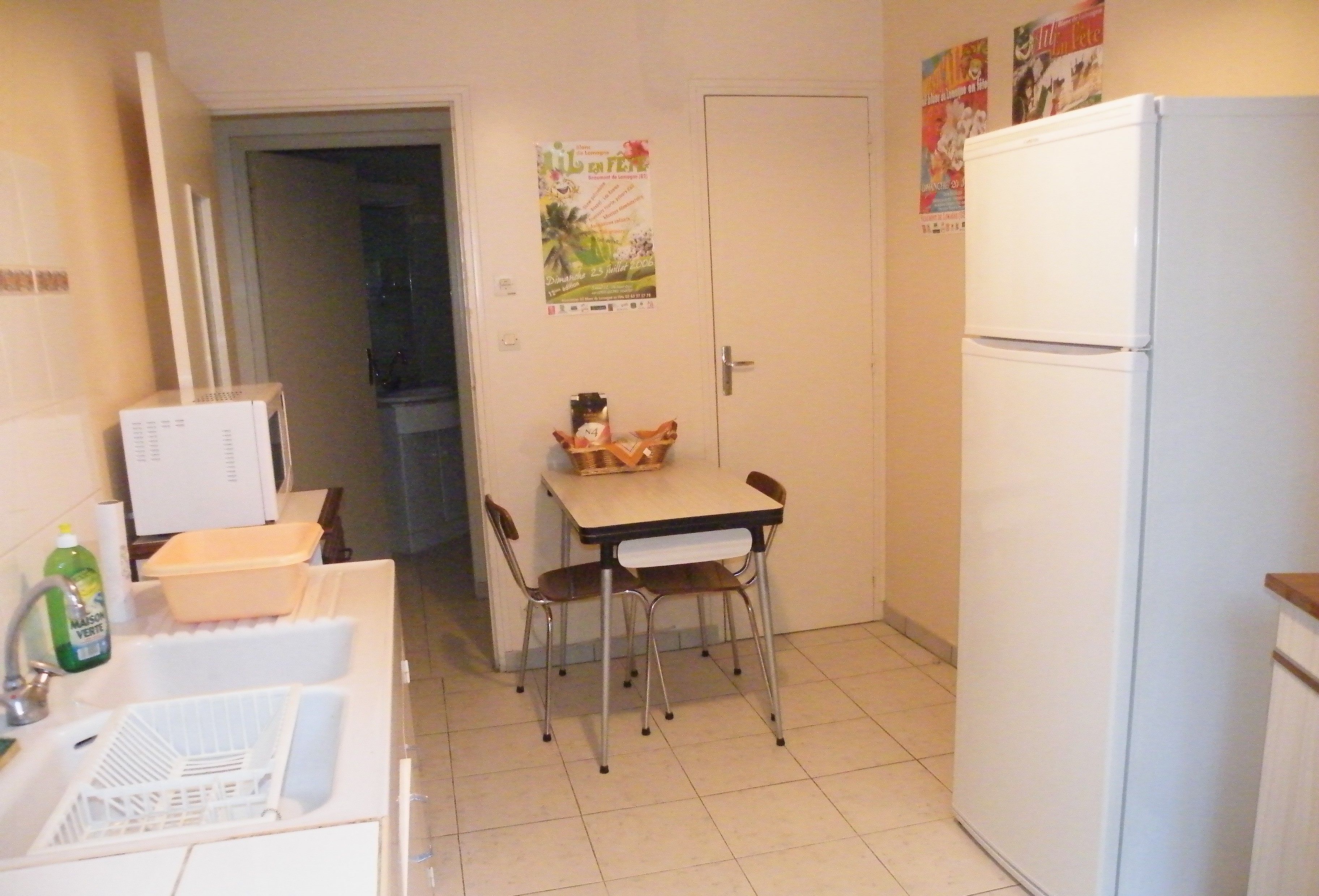 Una cocina pequeña con un refrigerador blanco, una mesa de comedor con sillas y un microondas. Hay una puerta que da a otra habitación y un cartel en la pared.