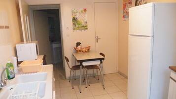 Una cocina pequeña con un refrigerador blanco, una mesa de comedor con sillas y un microondas. Hay una puerta que da a otra habitación y un cartel en la pared.