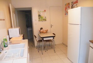Een kleine keuken met een witte koelkast, een eettafel met stoelen, en een magnetron. Er is een deur naar een andere kamer en een poster aan de muur.
