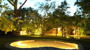 A circular fire pit surrounded by stone edging, set in a grassy area with trees and a wooden structure in the background.
