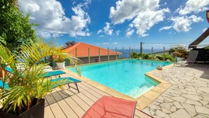 A swimming pool with a wooden deck, lounge chairs, and a red-roofed building in the background. - Paradyse-conditioned luxury villa, Saint-luce Martinique Pool (Sainte Luce)