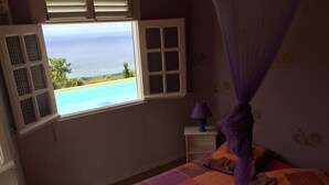 A bedroom with a bed, a window with a view of the ocean, a nightstand with a lamp, and a purple canopy. - Paradyse-conditioned luxury villa, Saint-luce Martinique Pool (Sainte Luce)