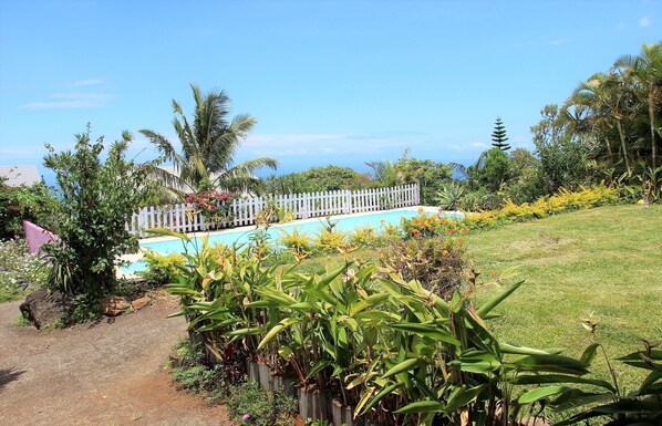 Una piscina rodeada por una cerca blanca, abundante vegetación y un cielo despejado.