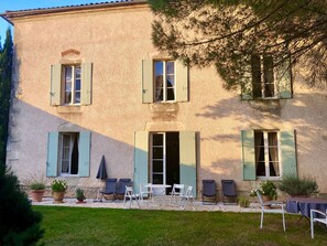Un edificio de dos pisos con fachada de piedra, varias ventanas con contraventanas y un patio con mobiliario exterior.