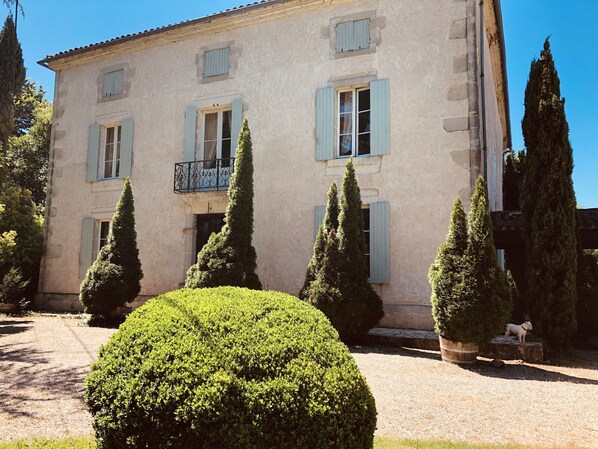 Un edificio de dos pisos con contraventanas azul claro, un balcón con barandilla de hierro forjado y un jardín bien cuidado con setos recortados y un arbusto redondo.