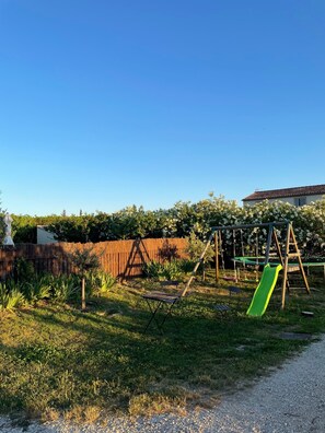 Ein Spielplatz mit Rutsche und Schaukeln, umgeben von einem Zaun und viel Grün.
