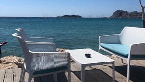 A wooden deck with white metal chairs and tables, overlooking a calm sea with sailboats and a distant island.
