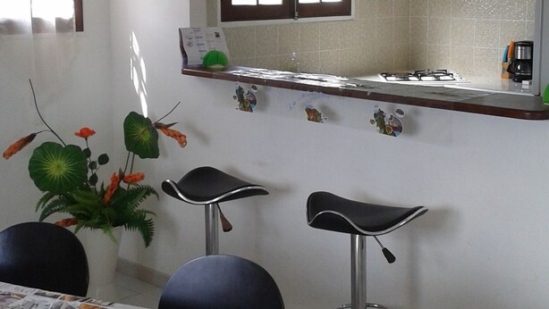 Een kleine, schone kamer met een bed, een tafel met een menukaart, twee stoelen, een raam met zonwering, en een kitchenette met een fornuis en koffiezetapparaat.