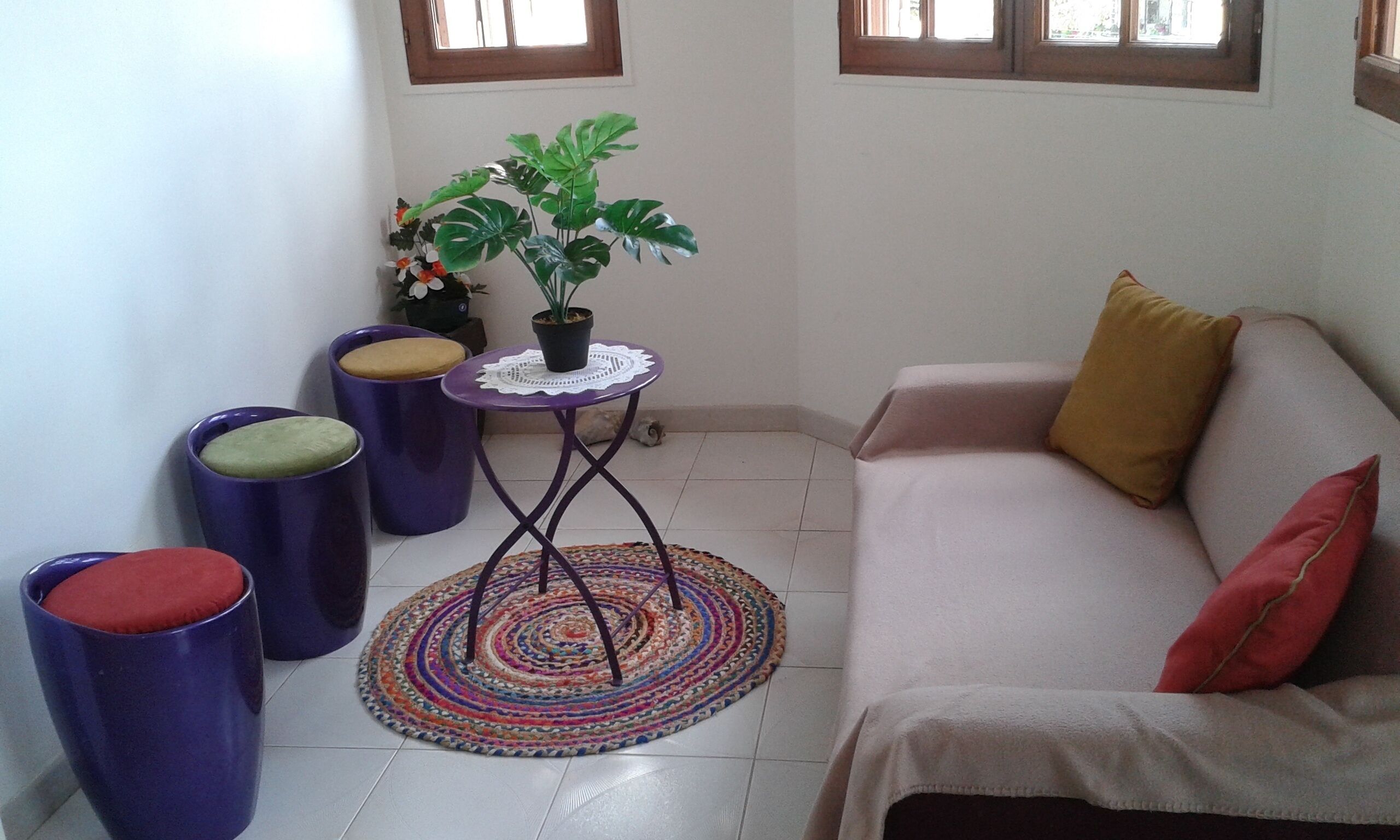 Un pequeño espacio de estar con un sofá, dos taburetes, una mesita con una planta y un tapete colorido.