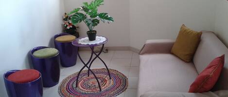 Un petit espace de vie comprenant un canapé, deux tabourets, une petite table avec une plante et un tapis coloré.