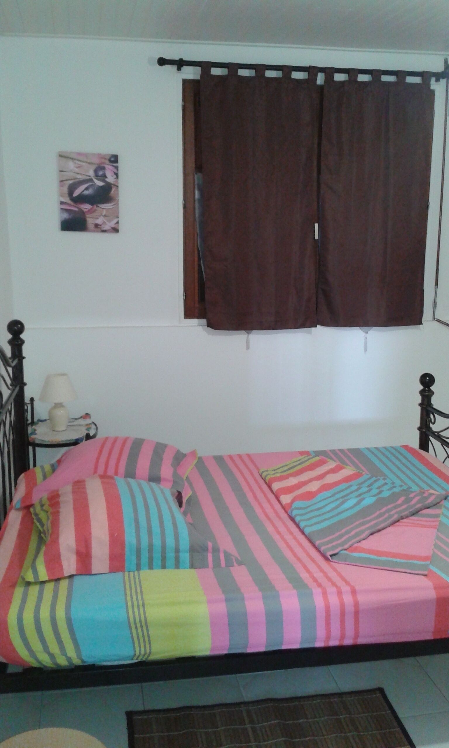 A single bed with a colorful striped bedspread, a bedside table with a lamp, and a window with brown curtains.