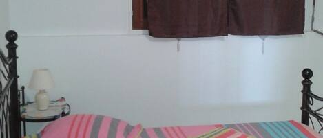 A single bed with a colorful striped bedspread, a bedside table with a lamp, and a window with brown curtains.