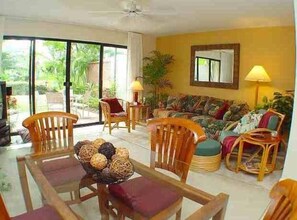 A spacious living room with a large sofa, wooden chairs, and a dining table with a centerpiece. - 1BR/2BA Maui Kamaole/Wailea Border. K Cluster @ Tropical Pool! (Kihei)
