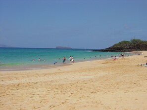 A sandy beach with clear turquoise water, a rocky outcrop in the distance, and people enjoying the beach. - 1BR/2BA Maui Kamaole/Wailea Border. K Cluster @ Tropical Pool! (Kihei)