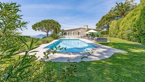 Area piscina privata con vista libera su una casa sullo sfondo, circondata da vegetazione rigogliosa e un prato curato.