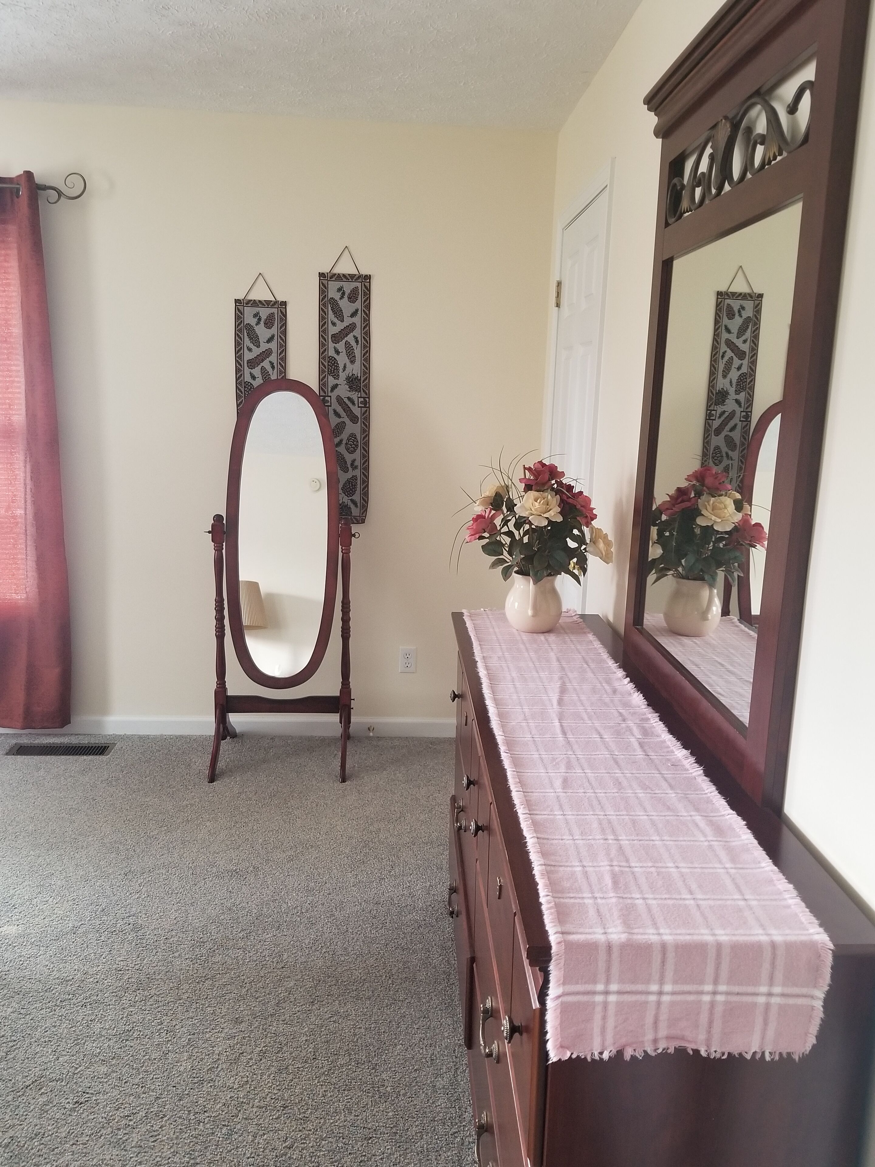 Une chambre à coucher avec une commode en bois, une coiffeuse dotée d’un miroir ovale, un vase rempli de fleurs et des éléments décoratifs accrochés au mur.