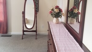 Un dormitorio con un aparador de madera, una tocador con un espejo ovalado, un florero con flores y adornos colgantes en la pared.