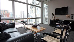 A modern office space with a large window, a black leather sofa, a wooden table, and a black desk with a flat-screen TV.