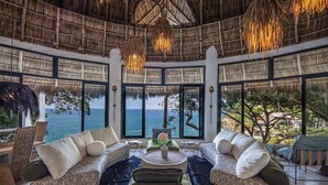 A spacious room with a large window offering a view of the sea, a thatched ceiling, and a central seating area with a coffee table. - Villa Violeta Luxurious Secluded Beachfront Hideaway. Weekly & Monthly discounts (San Francisco)