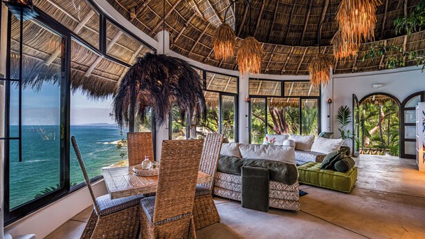 A spacious room with a large window overlooking the ocean, a thatched roof, wicker furniture, and a green sofa. - Villa Violeta Luxurious Secluded Beachfront Hideaway. Weekly & Monthly discounts (San Francisco)