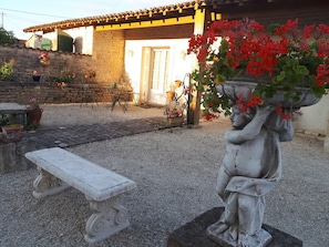 A courtyard with a stone bench, a statue holding a plant, and a rustic building with a tiled roof.