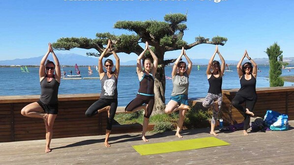 A group of people performing yoga on a wooden deck by a lake with a clear view of the water and distant mountains. - STUDIO 3/4P WITH CLIM, TERRACE 10M FROM THE LAC, BARCARES, CAP COUDALERE (Le Barcarès)
