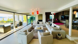 Een moderne woonkamer met een grote zithoek, een salontafel en een televisie. Er zijn ramen die uitzicht bieden op het landschap.