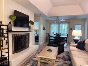 Una sala de estar con chimenea, televisor montado encima y comedor con mesa y sillas, además de ventilador de techo.