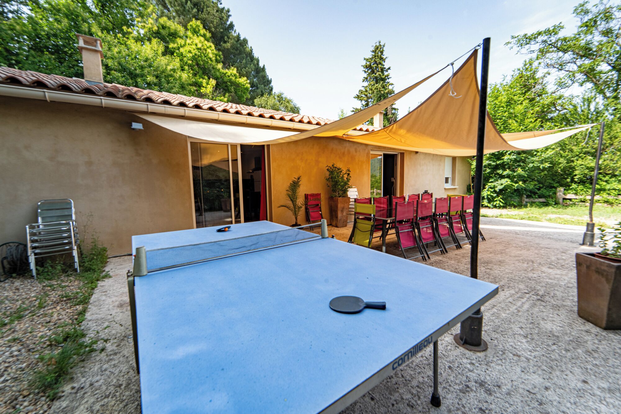 Een buitenpingpongtafel met een zonnescherm, omringd door stoelen en een gebouw met een pannendak.