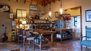 A rustic kitchen with wooden beams, a dining area with wooden chairs, and a variety of kitchenware and decorations on shelves.