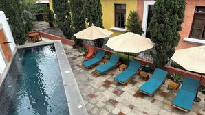 A swimming pool with lounge chairs and umbrellas, surrounded by trees and a building with a tiled patio. - Cozy Studio In Quiet Condo With Heated Pool (Antigua Guatemala)