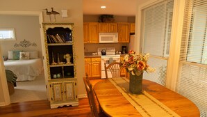 A dining area with a wooden table, chairs, and a vase of flowers. There is a kitchen in the background with wooden cabinets and a microwave. - Marsh-view Cottage in the Woods: surrounded by majestic oaks, birdsong & marsh! (Ridgeland)